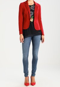 Roter taillierter Blazer, schwarzes Blumentop, schmale blaue Jeans und rote High Heels. Glatte Stoffe mit langen Ärmeln und taillierter Silhouette.