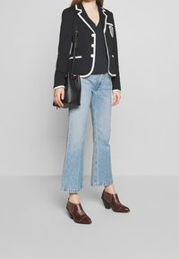 Femme portant un blazer noir avec des bordures blanches, un top noir, un jean cropped bleu clair, des chaussures à talons marron et tenant un sac à bandoulière noir.