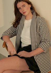 Blazer s kostkovaným vzorem v černobílé barvě s ohrnutými rukávy, oblečený přes bílý pletený svetr a černou sukni, sedící na dřevěné židli.