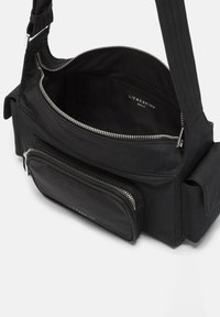 Sac à épaule en tissu noir avec un compartiment principal zippé, une poche externe et une sangle réglable. L'intérieur présente une doublure lisse.