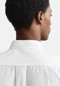 Camicia bianca con colletto strutturato, presenta un inserto posteriore e un bottone vicino al colletto, realizzata in tessuto di cotone liscio.