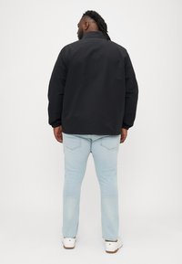 Crna, lagana jakna s uspravnim ovratnikom, elastičnim manžetama i opuštenim krojem, uparena sa svijetloplavim trapericama i bijelim cipelama.