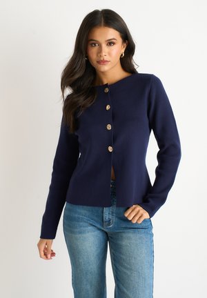 Young woman with long dark hair wearing a navy blue buttoned cardigan and light blue jeans, standing against a plain white background.