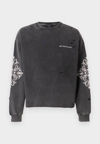 Sudadera desgastada de color gris oscuro con patrones decorativos en blanco en las mangas y el texto "NO DESPERDICIES CULTURA" en el pecho.