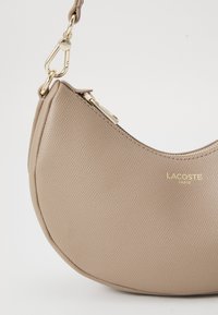 Beige textured leather crescent handbag with gold zipper and clasp, featuring Lacoste Paris logo in gold lettering.
