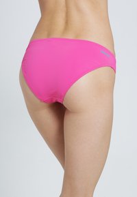 Rosa Bikini-Unterteil mit glatter Textur, niedrigem Schnitt und kontrastierendem blauen Logo auf der Rückseite. Gestickte Kanten sind sichtbar.