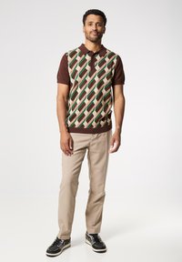 Camisa polo de manga corta con un patrón geométrico en verde, marrón y beige; combinada con pantalones beiges y zapatillas negras.