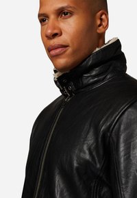 Zwarte leren jas met hoge kraag, voorzien van een ritssluiting en beige fake fur voering langs de kraag en voorkant, met een gestructureerde afwerking.