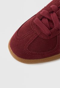 Bordó semišová teniska s texturovaným povrchem, gumovou podrážkou a plochými tkaničkami. Má zaoblenou špičku a prošití po stranách.