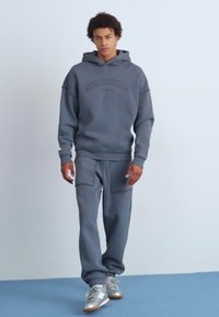 Szary hoodie z pasującymi szarymi dresami. Miękki materiał, luźny krój, przednie kieszenie i kaptur ze sznurkiem. Model nosi srebrne sneakersy.