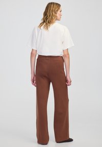 Bruine wijde broek met een gladde textuur en een elastische tailleband, gecombineerd met een witte top met korte mouwen. Gezien vanuit de achterkant.