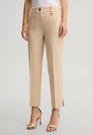 Frau trägt beige, maßgeschneiderte, knöchellange Hosen mit seitlichen Schlitzen, kombiniert mit goldenen High-Heel-Sandalen vor einfarbigem Hintergrund.