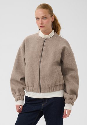 Joven mujer con cabello rubio recogido en una coleta lleva una chaqueta beige con cremallera sobre un jersey de cuello alto blanco y vaqueros azul oscuro, de pie frente a un fondo liso.