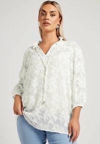 Blusa bianca trasparente con ricamo floreale, scollatura a V e dettaglio con coulisse. Presenta maniche a sbuffo e una vestibilità ampia.