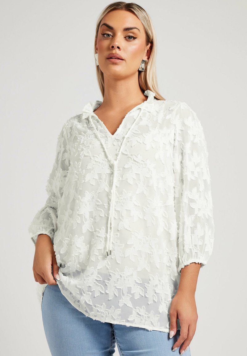 Blusa bianca trasparente con ricamo floreale, scollatura a V e dettaglio con coulisse. Presenta maniche a sbuffo e una vestibilità ampia.