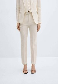 Costume en lin beige composé d'une veste cintrée, d'un gilet assorti et d'un pantalon slim. Accompagné de sandales à brides avec un détail subtil de boucle.