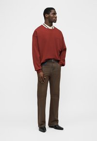 Jeune homme debout, portant un sweat-shirt oversize couleur rouille rouge par-dessus une chemise blanche à col, un pantalon marron à jambes droites et des chaussures noires pointues.