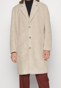 Uomo che indossa un cappotto di shearling beige fino al ginocchio con rever e tre bottoni, abbinato a un top nero e pantaloni color ruggine.