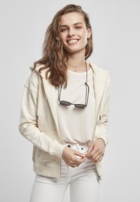 Kremfarget zip-hettegenser med kengurulomme, bæres over en lys t-skjorte og hvite jeans. Har hette og solbriller hengende fra nakken.