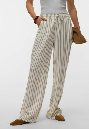 Persoon die losse beige broek met verticale grijze strepen draagt, een grijs mouwloos topje, bruine instapschoenen en een bruine clutch vasthoudt.