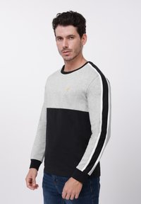 Siva i crna majica dugih rukava s okruglim izrezom, s donjim dijelom u čvrstoj crnoj boji i bijelim bočnim prugama. Uočljiv detalj žutog logotipa.