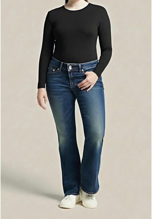 Personne portant un haut à manches longues noir, un jean bleu en denim et des chaussures blanches, debout devant un fond beige neutre.