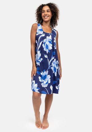 Vrouw glimlachend, staand op blote voeten, dragend een mouwloze navy blauwe jurk met groot wit en blauw bloemenpatroon.