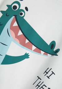 Camiseta blanca con un cocodrilo de dibujos animados que tiene una gran sonrisa, en colores verde y azul, y un texto divertido que dice "HOLA" en negro.
