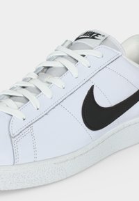 Άσπρο χαμηλό sneaker της Nike με μαύρο λογότυπο swoosh, λευκά κορδόνια, υφασμένο πέλμα και επενδεδυμένο περιμετρικά μέρος, πάνω σε απλό φόντο.