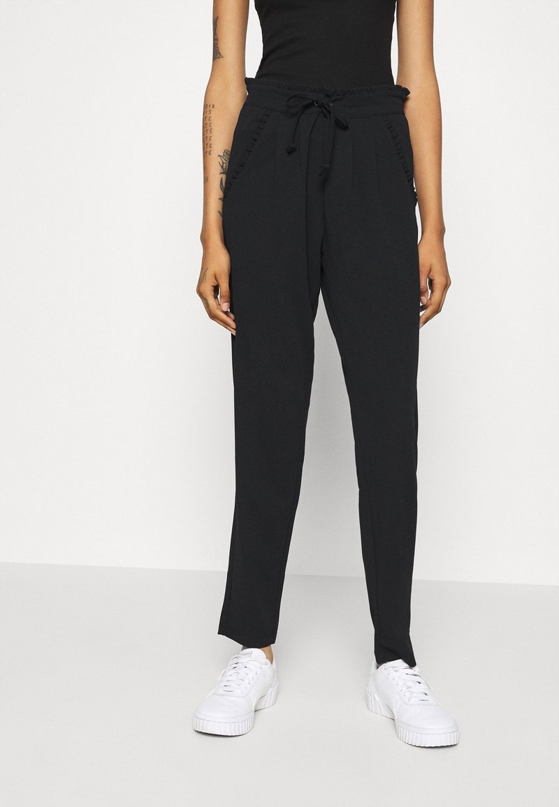 Pantalones negros con cordón en la cintura, con una banda texturizada y bolsillos laterales, combinados con zapatillas blancas. Tela suave, diseño de pierna recta.