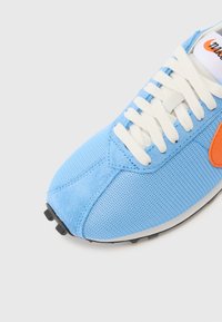 Sneaker in rete blu e suede con lacci bianchi, presenta un logo laterale arancione e una suola in gomma nera. Pannelli testurizzati e cuciture visibili.