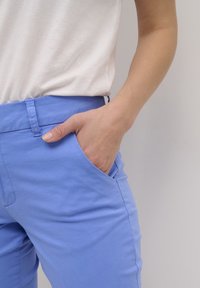 Pantalon bleu clair en mélange de coton, avec un devant plat, des poches latérales et une coupe ajustée. Porté avec un haut blanc.