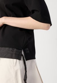 Black short-sleeve top with satin accent at the hem, featuring a logo patch on the left side. Textured and structured fabric.