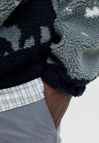 Ręka w kieszeni, na sobie szare spodnie, koszula w kratkę i polarowa kurtka w ciemno- i jasnoszarym wzorze.