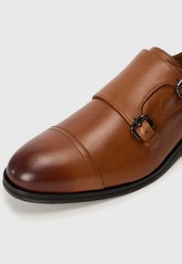 Zapato de vestir de cuero marrón con doble hebilla, puntera redondeada y acabado suave. Presenta una puntera chapada cosida y una suela negra.