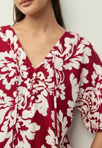 Blouse à imprimé floral rouge et blanc avec un décolleté en V et un détail à lacet. Manches courtes et coupe ample dans un tissu texturé.