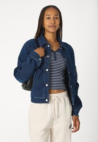 Giacca di jeans blu con bottoni argentati, vestibilità rilassata, indossata sopra un top a righe blu navy e bianche. Pantaloni felpati beige con coulisse e borsa nera.