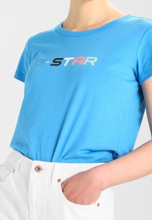 Femme portant un t-shirt bleu vif avec le texte "G-STAR" et un pantalon blanc taille haute, la main droite dans la poche.