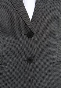 Gros plan d'un blazer texturé gris foncé avec deux boutons noirs fermés et une chemise blanche visible sous le col.