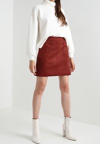 Weißes, strukturiertes Langarmoberteil, kombiniert mit einem roten Wildleder-Minirock mit Fronttaschen und weißen Knöchelboots. Einfaches und modernes Design.