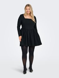 Smilende kvinne iført svart langermet kjole med firkantet halslinning, svarte strømpebukser og svarte spisse flate sko, stående på lys grå bakgrunn.