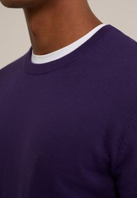 Lila stickad tröja med rund halsringning, som har en texturerad yta och ribbade detaljer vid kragen och ärmarna.