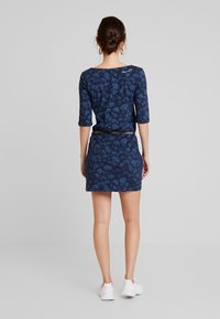 Kvinna med håret i knut, iklädd en blå blommig klänning med trekvartsärmar, brun bälte och vita sneakers, står med ryggen mot kameran på en grå bakgrund.