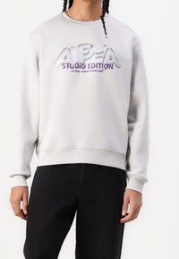 Jasnoszara bluza z okrągłym dekoltem, ściągaczami przy mankietach i u dołu. Posiada wyraźne, odblaskowe logo "ALPHA STUDIO EDITION" w kolorze purpurowym.