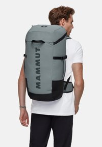 Grå Mammut-ryggsäck med svarta accenter, stor cylindrisk form, vadderade remmar och en djärv logotyp på sidan. Texturerad tyg.