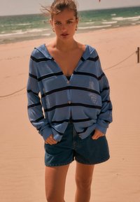 Lichtblauw gestreept overhemd met knopen en een losse pasvorm en ribbelstructuur, gecombineerd met donkerblauwe spijkerbroekshorts. Strandachtergrond.