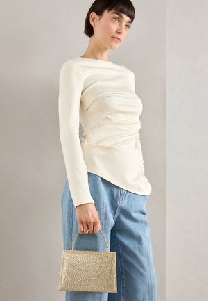 Femme aux cheveux courts et foncés portant un haut à manches longues écru et un jean bleu, tenant un petit sac à main doré texturé par la poignée.
