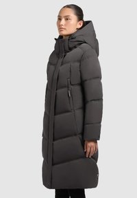 Sieviete, kura valkā garu melnu dūnu ziemas mēteli ar augstu apkakli un kapuci, stāv sānis pret vienkāršu fonu.