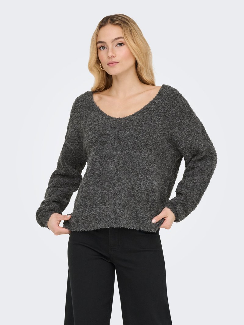 Femme blonde portant un pull gris foncé à manches longues et duveteux ainsi qu'un pantalon noir, tenant l'ourlet du pull avec ses deux mains.