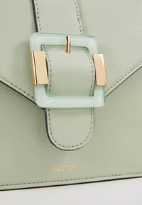 Světlá zelená kožená kabelka se čtvercovou mátovou sponou a zlatými kovovými detaily. Jsou viditelné čisté linie a detailní prošívání. Logo ALDO je viditelné.